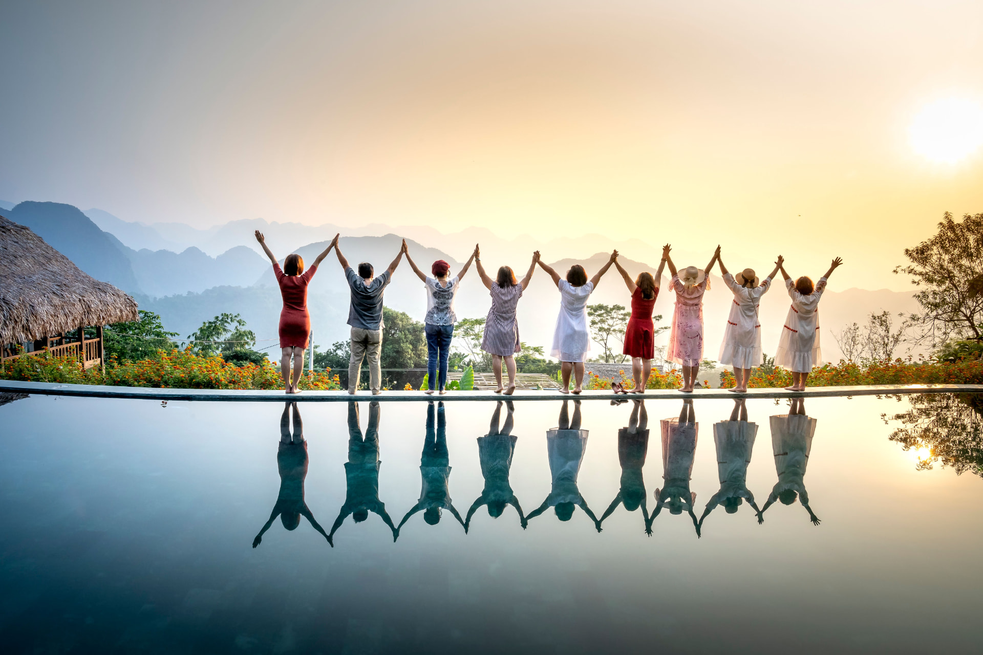 A group of friends with raised hands beside an infinity pool at sunset